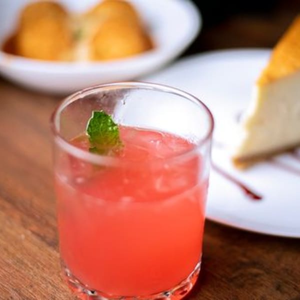 A vibrant red cocktail with a mint garnish, cheesecake, and arancini on a wooden table.
