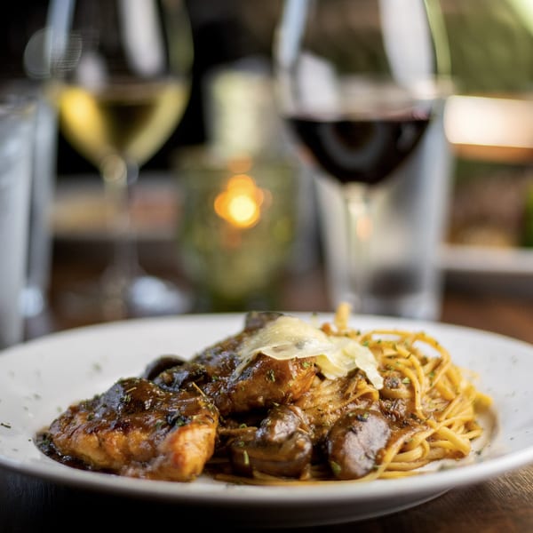 Pignetti's chicken and pasta dish with mushrooms and shaved cheese, with wine glasses in the background.