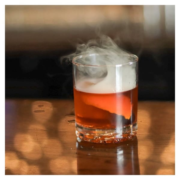 A smoking Pignetti's cocktail, a red drink in a clear glass, sits on a wooden surface.