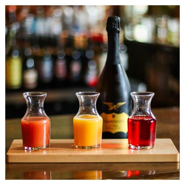Three carafes of colorful juices and a bottle of champagne on a wooden board.