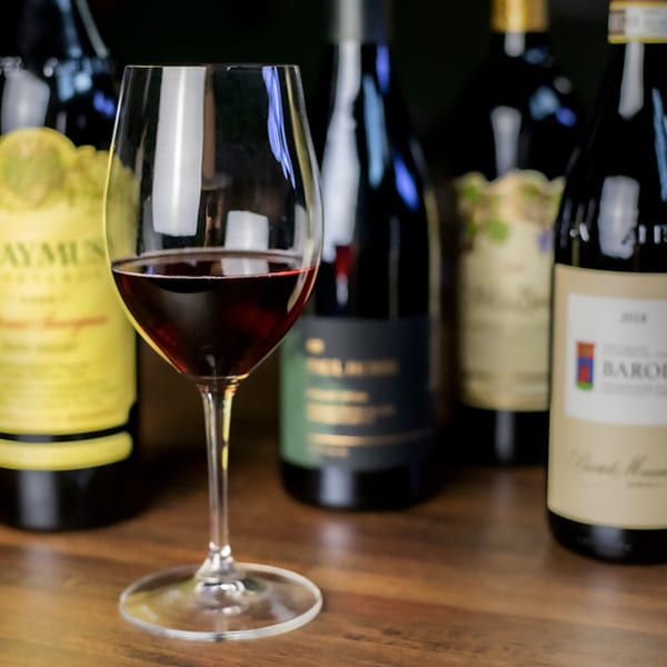 A wine glass filled with red wine sits on a wooden table, surrounded by several wine bottles.