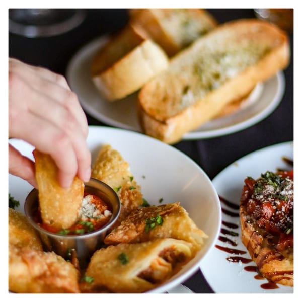 Hand dipping a fried ravioli into marinara sauce, with garlic bread and bruschetta in the background.