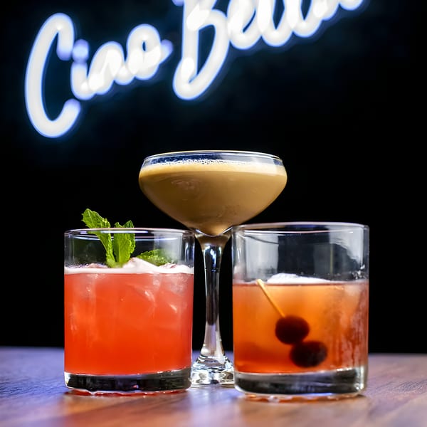 Three Pignetti's cocktails, one pink with mint, one brown, and one amber with cherries, under a neon "Ciao Bella" sign.
