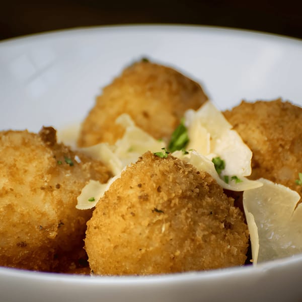 Close-up of golden-brown arancini balls topped with shaved Parmesan and fresh herbs in a white bowl.
