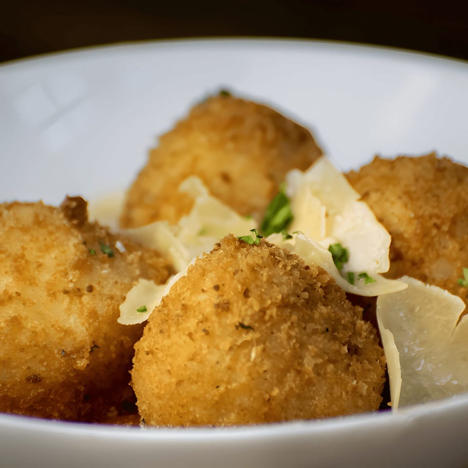 Close-up of golden-brown arancini balls topped with shaved Parmesan and fresh herbs in a white bowl.