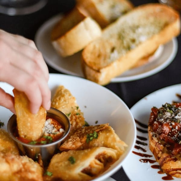 Hand dipping a fried Italian appetizer into a small metal cup of red sauce with garlic bread.