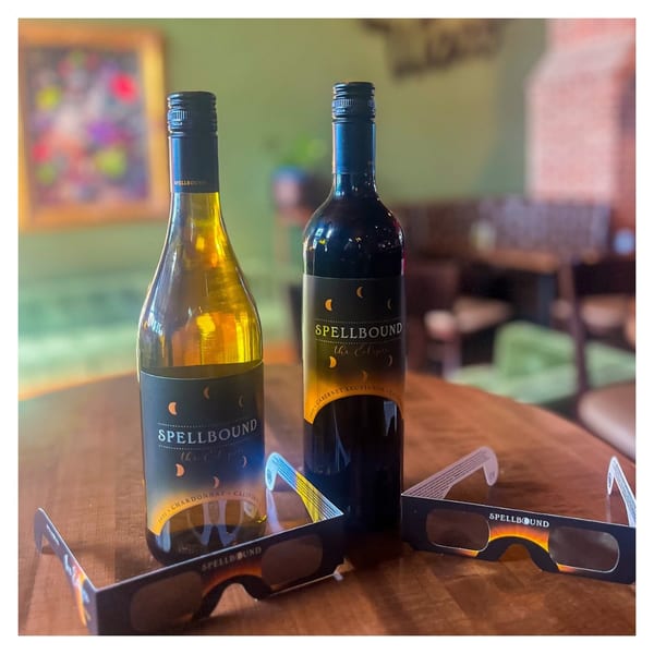 Two Spellbound wine bottles, one white and one red, with eclipse glasses on a wooden table.